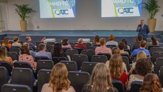 Foto, em primeiro plano, do público interno assistindo à abertura da apresentação. Há várias pessoas, de costas, sentadas em poltronas azuis do auditório do TCE/SC. À frente delas, o conselheiro Aderson Flores, falando ao microfone. Ele veste terno e gravata e usa óculos de grau. Ao fundo, no telão, é projetada uma apresentação com logo do MMD-TC.