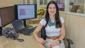 Foto da auditora fiscal de controle externo Juliana Medeiros das Neves Moser. Ela é uma mulher branca, de cabelos compridos e castanhos. Veste uma blusa branca e calça bege. Está sentada em uma cadeira e sorri. Atrás dela, um monitor mostra o logo do TCE/SC. 