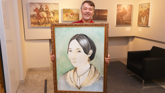Foto do servidor do TCE/SC Rogerio Guilherme de Oliveira segurando uma quadro com o desenho em aquarela da Anita Garibaldi - uma mulher branca, com cabelos escuros presos num coque. Atrás dele, vários quadros com a temática de Anita Garibaldi pendurados nas paredes.