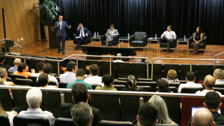 Imagem mostra os palestrantes no palco e a plateia em primeiro plano. À esquerda, no alto, falando ao microfone, está o diretor da Ouvidoria do TCE, Paulo César Salum