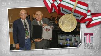 Banner com fundo em tons de cinza. Sobre o fundo, foto do ministro Jorge Mussi com a Medalha do Mérito Tribunal de Contas e com o diploma, em pé, junto com o conselheiro do TCE/SC Wilson Wan-Dall. No canto superior direito, imagem da medalha. No canto inferior direito, imagem da logomarca do TCE/SC.