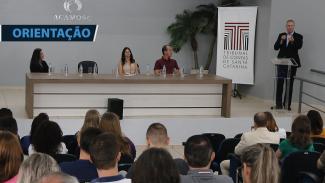 Foto da bancada de autoridades durante o evento na Acamosc. Entre eles, a presidente da Associação, Franciéli Werlang. No púlpito, à direita, o presidente Herneus De Nadal fala ao público. À sua frente, pessoas assistem à sua fala, sentadas em cadeiras.