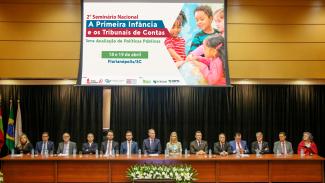 Imagem mostra a mesa de abertura do seminário da primeira infância. No centro da imagem, acima das pessoas, telão mostra o cartaz do evento, onde aparecem crianças do lado direito da tela e a descrição do seminário no lado esquerdo.