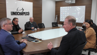 Foto horizontal com sete pessoas sentadas em volta de uma mesa, entre elas, o conselheiro Herneus De Nadal e uma mulher. Ao fundo, a identificação da Unochapecó