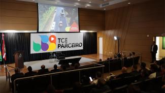 Foto do palco do auditório do TCE/SC. No palco, sobre  uma cortina presta ao fundo está a logomarca do TCE Parceiro (em fundo branco, 5 formas coloridas que parecem gotas, verde, roxo, azul amarelo e vermelho). Acima, o telão exibe imagem de estrada em obras com caminhões