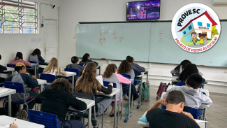 Imagem mostra crianças de costas em uma sala de aula de escola visitada. No fundo há um quadro e, acima dele, um monitor/televisor. À esquerda, uma janela basculante está aberta. No lado superior direito, há um selo do Programa de visita às escolas, que é um desenho de um prédio branco com a inscrição “escola” e duas crianças, um menino branco e uma menina preta, à frente