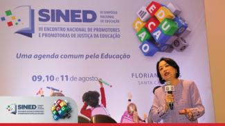 Imagem mostra palestrante falando ao microfone em primeiro plano. Ao fundo, a projeção com a logomarca do evento.
