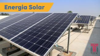 Imagem mostra em primeiro plano placas de coleta de energia solar, os painéis fotovoltaicos. No alto, à esquerda, sobre fundo amarelo, está escrito em azul a expressão energia solar.