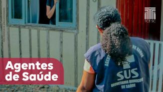 Imagem mostra foto de um agente de saúde em visita a uma casa. A gente aparece à direita na foto, de cabelo amarrado, vestindo um colete azul escuro onde se pode ler, em letras brancas, a identificação como agente e o logo do SUS. Ao fundo, em uma janela azul claro numa parede de madeira, aparece uma pessoa sem identificação