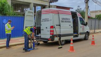 Imagem mostra van do TCE centralizada. À esquerda, um técnico do tribunal, vestindo colete amarelo, registra em imagens o trabalho de outro técnico, que com uma máquina perfuradora retira um pedaço do asfalto. 