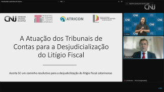 Banner com a imagem do evento captada do vídeo. Na tela, o título da palestra dos integrantes do TCE/SC — “A atuação dos Tribunais de Contas para a desjudicialização do litígio fiscal” e, no canto direito, de cima para baixo, o logo do CNJ, e as imagens da intérprete de libras, do conselheiro José Nei Ascari e de um marcador de tempo, com registro de nove minutos e um segundo.