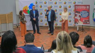 Foto de quatro pessoas em pé durante a primeira etapa do evento sobre ouvidorias: dois homens e duas mulheres. Entre eles, o conselheiro Wilson Wan-Dall e o diretor da Ouvidoria do TCE/SC, Paulo Cesar Salum. Eles estão um ao lado do outro. O conselheiro fala ao microfone. Atrás deles, há um banner com vários logos da Amunesc e, ao lado, um banner com informações da ouvidoria do TCE/SC. À frente, de costas para a foto, há pessoas que assistem à fala. 