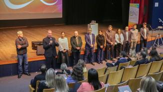 Foto do presidente Herneus De Nadal falando ao público durante etapa de São José do Ciclo de Estudos. Ele está em pé, em um auditório, em frente ao palco, acompanhado de outras dez pessoas, que estão ao seu lado. À sua frente e de costas para a foto, estão as pessoas que assistem à sua fala. Todas estão sentadas em cadeiras. 