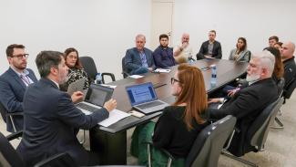 Foto de uma das reuniões do Plano de Correição 2023. Há 14 pessoas sentadas ao redor de uma mesa. Sobre a mesa, há notebooks, garrafas, papéis e canetas.    