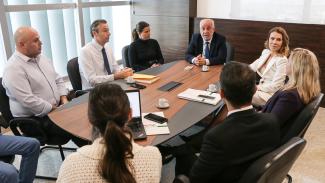 Reunião ocorre ao redor de uma mesa marrom e preta. Na foto aparecem de frente três homens e duas mulheres. Uma mulher aparece de lado. Outra mulher e um homem aparecem de costa. Sobre a mesa há um laptop aberto, algumas xícaras de café e telefones celulares.