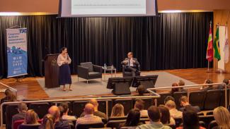 Foto do palco do auditório do TCE/SC durante o painel sobre controle da administração pública. Falando ao público, em pé e à esquerda, está a professora Jéssica Mota. À direita, sentado em poltrona, está o professor Rodrigo Valgas. Ao fundo, no telão, há um PowerPoint com apresentação da palestrante. 