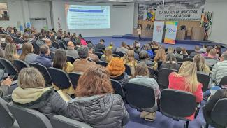 Foto horizontal dos participantes — homens e mulheres — do Ciclo de Estudos. Eles estão sentados em poltronas e de costas. Ao fundo, de frente, uma mulher, que está falando ao microfone. Na parede, ao fundo, a logo da Câmara Municipal de Caçador. Na parede lateral à esquerda (da foto), um slide sobre pesquisa de preço.