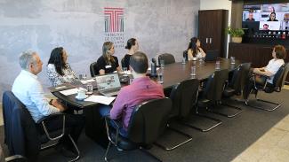 Foto da reunião dos integrantes do Projeto Lume. À esquerda, há sete pessoas sentadas ao redor de uma mesa, enquanto conversam com outras pessoas em telechamada, exibida na televisão, à direita da foto. Entre os presentes, o conselheiro substituto Gerson dos Santos Sicca e o chefe de gabinete Rafael Tachini.  