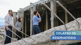 Foto do reservatório - obra em concreto - rompido. Na imagem, 2 homens e 1 mulher observam a estrutura danificada. No canto inferior direito, sobre um fundo azul está escrito "Colapso de Reservatório", em letras brancas.