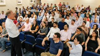 Presidente do TCE/SC, Herneus De Nadal, está em pé, à esquerda, falando ao microfone. Demais pessoas estão sentadas à sua frente.  Há cerca de 60 pessoas na imagem.