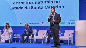 Foto do auditor da DAE do TCE/SC Osvaldo Faria de Oliveira. Ele, um homem branco de terno escuro, está ao lado esquerdo da foto. Atrás dele, no palco, um homem e uma muher estão sentado. Ao fundo um telão com uma imagem onde está escrito "desastres naturais no Estado de Santa Catarina.