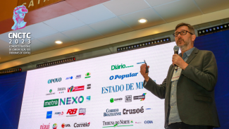 Banner com a imagem do palestrante, em pé, em primeiro plano e à direita. Ao fundo, painel de led com projeção de diversos nomes de veículos de imprensa. No canto superior esquerdo, o logotipo do evento. 