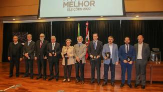 Dez pessoas, entre autoridades e representantes da NSC Comunicação, estão em pé e segurando uma publicação com capa preta. Atrás delas, há uma mesa, e, ao centro, as bandeiras do Brasil e de Santa Catarina e um banner do projeto. Acima, no telão, há o texto SC Ainda Melhor - Eleições 2022.
