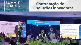 Banner com a imagem do servidor Jairo Wensing durante palestra no Summit Cidades 2022. Ele veste um terno cinza e está segurando um microfone em uma das mãos. Na foto, há mais pessoas, que assistem à sua apresentação.  Acima, o logotipo do evento, composto do referido nome em fonte branca. Ao lado direito, o texto “Contratação de soluções inovadoras”, em fonte cinza, sobre fundo branco. 