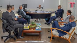 Foto de uma sala em que aparece uma mesa ao fundo, a frente da qual está sentado o presidente do TCE/PR, conselheiro Fernando Gumarães. Sobre a mesa há um monitor e materiais de escritório. Sentados defronte à mesa, em poltronas e cadeiras dispostas em duas filas de três, uma de cada lado da mesa, estão seis pessoas, entre elas o conselheiro Wilson Wan-Dall, que ocupa um lugar à direita, próximo à mesa. Ao centro há uma mesinha com uma folhagem e alguns livros. No canto superior direito, há a logo do TCE/SC