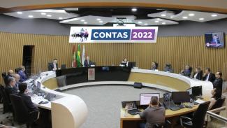 Foto mostra plenário do TCE/SC, que tem fundo amarelado. Na bancada, autoridades sentadas próximas a parede. No centro da imagem, uma inserção de banner menor com as palavras Contas 2022, em branco sobre fundo azul e roxo.  