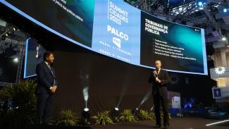 Imagem mostra um palco com um telão ao fundo. À esquerda, há um homem de terno escuro. À direita, também de terno escuro, está o presidente do TCE/SC, conselheiro Herneus de Nadal, que fala ao microfone. O telão mostra ao centro o logo do Summit Cidades. À direita, há legível a expressão Tribunal da Governança Pública.