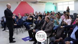   Imagem mostra o presidente do TCE/SC, Herneus De Nadal, em pé, à esquerda. Ele veste um traje preto. O restante da foto é tomado por pessoas sentadas em um auditório. No centro, parte inferior da imagem, há um selo escrito TCE/SC em Movimento, em fundo branco com letras pretas e setas vermelhas que indicam movimento