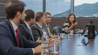 Banner horizontal com foto da comitiva do TCE-RJ reunidos com comitiva do TCE/SC diante de uma mesa enquanto conversam. Há quatro homens e uma mulher na foto, a assessora Flávia Ramos, do TCE/SC. Sobre a mesa, há copos de água.