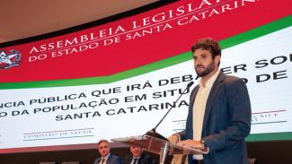 Foto do auditor fiscal Rafael Scherb, em frente ao púlpito, falando ao microfone. Ao fundo, painel luminoso com o banner identificando o nome do evento, e dois deputados sentados. 
