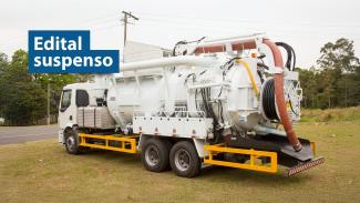 Banner horizontal com foto de um caminhão equipado com sistema de sucção e limpeza, com mangueiras e compartimentos acoplados na parte traseira, estacionado em área aberta com vegetação ao redor. No canto superior esquerdo, em tarjas azuis, o texto "Edital suspenso".