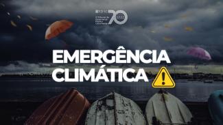 Banner horizontal com foto de paisagem costeira sob um céu escuro e carregado por nuvens de tempestade. No primeiro plano, há pequenos barcos virados de cabeça para baixo alinhados à beira da água. Aparecem guarda-chuvas coloridos sendo levados pelo vento e folhas espalhadas pelo ar, sugerindo condições climáticas extremas. Sobre a imagem, em fonte branca, o texto "Emergência climática", ao lado de ícone de atenção amarelo.