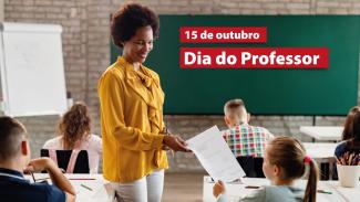 Foto de uma professora entregando uma folha de papel a uma criança sentada em uma carteira. A professora é uma mulher negra, de cabelos crespos, que sorri e usa roupa amarela. Há outras crianças sentadas em carteiras enfileiradas em uma sala de aula. Ao fundo, no quadro de giz, o texto “15 de outubro. Dia do Professor”, em destaque em retângulos vermelhos.