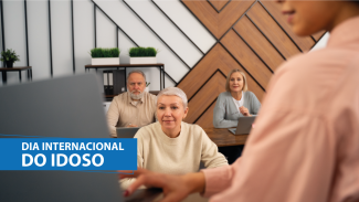 Banner horizontal com foto de três idosos sentados diante de mesas em uma sala, olhando para o que mostra uma mulher que está à frente deles, de costas para a foto, em primeiro plano e com desfoque. Sobre a imagem, na lateral esquerda, em tarja azul, o texto “Dia Internacional do Idoso”.