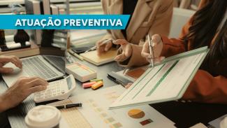 Imagem mostra uma mesa com pessoas trabalhando ao redor, sem aparecer os rostos. Uma pessoa manuseia uma calculadora, a outra mostra uma planilha e uma terceira está apontando para essa planilha. No alto, à esquerda, escrito em branco no fundo azul, está escrito Atuação Preventiva