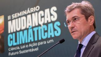 Foto do conselheiro José Nei Ascari discursando no púlpito. Ele é um homem branco, de cabelos grisalhos. Veste terno e gravata e usa óculos de grau. Ao fundo, banner com o título do evento, “segundo Seminário Mudanças Climáticas: ciência, lei e ação para um futuro sustentável”.