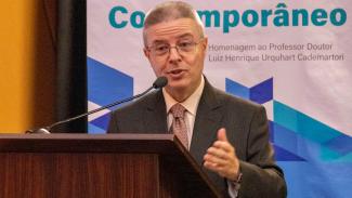 Foto do ministro do TCU Antonio Anastasia falando ao púlpito. Ele é um homem branco de cabelos grisalhos. Está vestindo terno preto, camisa branca e gravata estampada e usa óculos. Ao fundo, há o banner do Congresso.