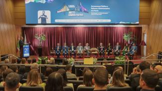 Foto horizontal que mostra, em primeiro plano, a plateia participante do evento, de costas, sentada em poltronas do auditório. Ao fundo, no palco, as autoridades lado a lado, durante a abertura do Congresso. Há 11 pessoas, lado a lado. Entre os presentes, o presidente do TCE/SC, Herneus De Nadal, o presidente do Idasc, José Cristóvam, e o homenageado, João Henrique Blasi. Presentes ainda, membros do MPTC/SC, do TJSC e da OAB/SC.