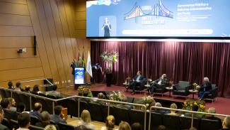Banner horizontal com foto que mostra parte da plateia, de costas, assistindo a um dos painéis do congresso. Há várias pessoas sentadas em poltronas do auditório, lado a lado. No palco, entre os presentes, os desembargadores Hélio Pereira e João Henrique Blasi, o conselheiro Adircélio Ferreira Júnior e o presidente do Idasc, José Cristóvam. Ao fundo, no telão, é exibida a identidade visual do evento.  