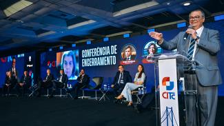 Foto do palco do IV CITC. À direita, no púlpito, o ministro do STF Flavio Dino discursa. Ao funo, telão com imgame da ministra Carmen Lucioa e os dizeres Conferencia de Encerramento. No palco ainda estão os presidentes do IRB, Atricon , TCE/SC r o ministro André Mendonça.