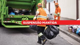 Banner horizontal com foto de trabalhadores da coleta de resíduos urbanos. Em primeiro plano, um funcionário uniformizado com roupa laranja e faixas refletivas carrega sacos de lixo em direção a um caminhão de coleta verde. Ao fundo, outro trabalhador sinaliza próximo ao veículo, enquanto sacos de resíduos aparecem dispostos na calçada. Sobre a imagem, uma faixa vermelha em destaque exibe o texto "Suspensão mantida". 