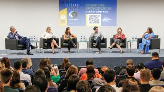 Foto do evento. Ao fundo, sobre o palco, o palestrante e os cinco debatedores, sendo quatro mulheres e um homem, sentados em poltronas pretas e de frente para a plateia, cujas pessoas também estão sentadas. 