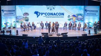Banner horizontal com foto do auditório lotado durante a realização do 9º Conacon – Congresso Nacional dos Auditores de Controle Externo dos Tribunais de Contas do Brasil. No palco, autoridades e participantes estão distribuídos em cadeiras ao fundo, diante de um grande painel com a identidade visual do evento. Ao centro, a auditora fiscal de controle externo do TCE/SC, Juliana Moser, recebe uma homenagem durante a cerimônia, acompanhada por representantes da organização. 