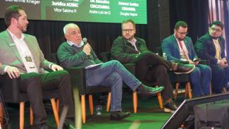 Imagem mostra cinco homens sentados em cadeiras em um palco. O segundo homem da esquerda para a direita, o conselheiro Luiz Eduardo Cherem, está falando ao microfone