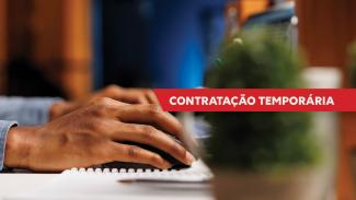 Imagem em ambiente interno mostrando mão apoiada sobre mouse de computador ao lado de um teclado, sobre a mesa. Sobre a imagem, há faixa vermelha com o texto “Contratação temporária”, em letras brancas. 
