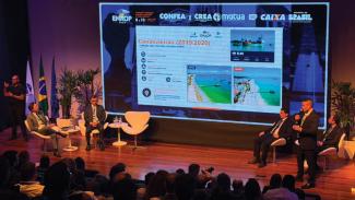 Imagem mostra um palco com pessoas nele. Ao fundo, há uma projeção em tela da apresentação da DLC.